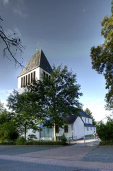 Kath. Kirche St. Heinrich 1 Ein moderner Kirchturm mit schwarzen Schindeln ragt über die Bäume, flankiert von blauem Himmel.