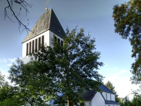 Kath. Kirche St. Heinrich 1 Ein moderner Kirchturm mit schwarzen Schindeln ragt über die Bäume, flankiert von blauem Himmel.