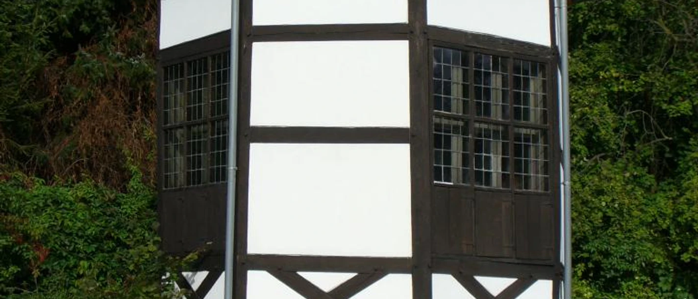 Fachwerkhaus mit achteckigem Grundriss und ziegelbedecktem Dach, umgeben von Bäumen und Sträuchern.