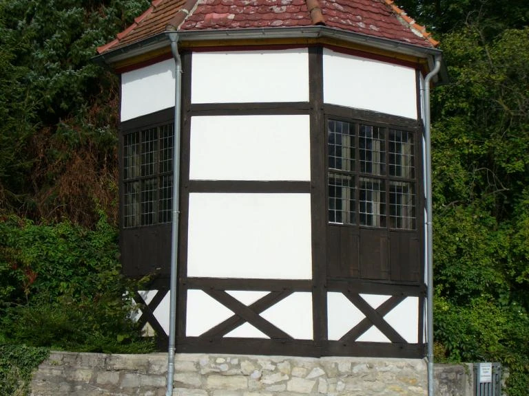 Fachwerkhaus mit achteckigem Grundriss und ziegelbedecktem Dach, umgeben von Bäumen und Sträuchern.