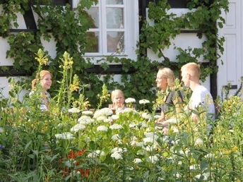 Eine Familie sitzt inmitten blühender Pflanzen vor einem Fachwerkhaus mit Efeubewuchs.
