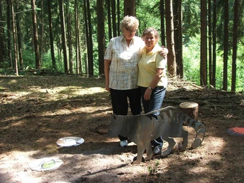 Inge Spangenberg, Margaretha Zantow Zwei Frauen stehen lächelnd im Wald auf einem Pfad neben einer skulpturalen Darstellung eines Wolfes.