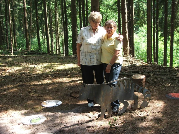 Inge Spangenberg, Margaretha Zantow Zwei Frauen stehen lächelnd im Wald auf einem Pfad neben einer skulpturalen Darstellung eines Wolfes.