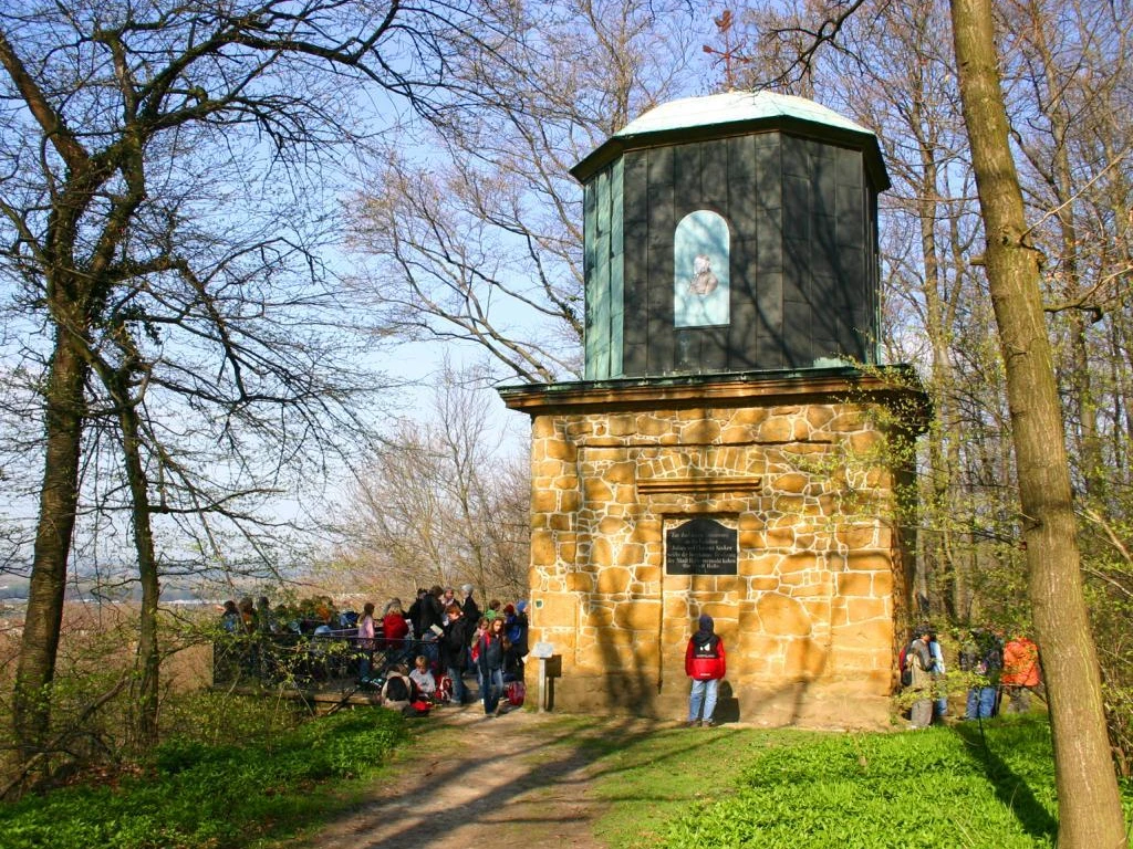 Eine Gruppe von Menschen steht vor einem steinernen Aussichtsturm im sonnigen Wald.