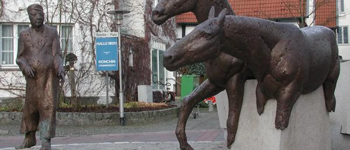 Halle Westfalen, Denkmal "Haller Willem" Zwei bronzene Pferdeskulpturen springen über Steinmauern, während ein Mann in Mantel sie beobachtet.