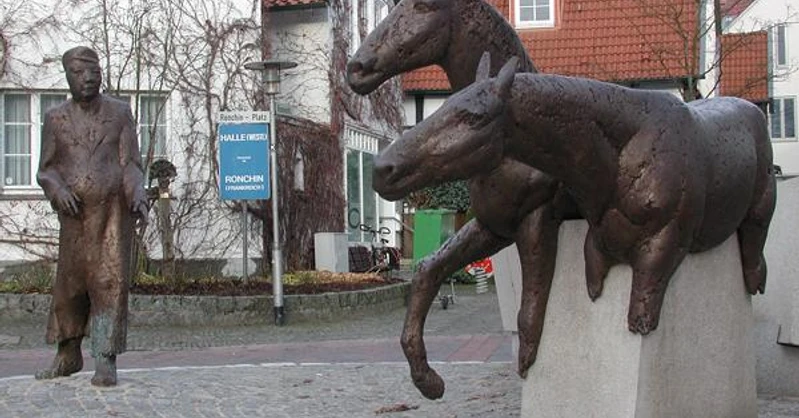 Halle Westfalen, Denkmal "Haller Willem" Zwei bronzene Pferdeskulpturen springen über Steinmauern, während ein Mann in Mantel sie beobachtet.