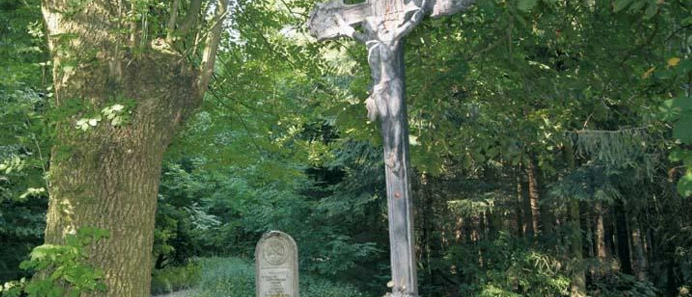 Kruzifix im Lippspringer Wald, umgeben von dichten Bäumen, mit einem Grabstein im Hintergrund.