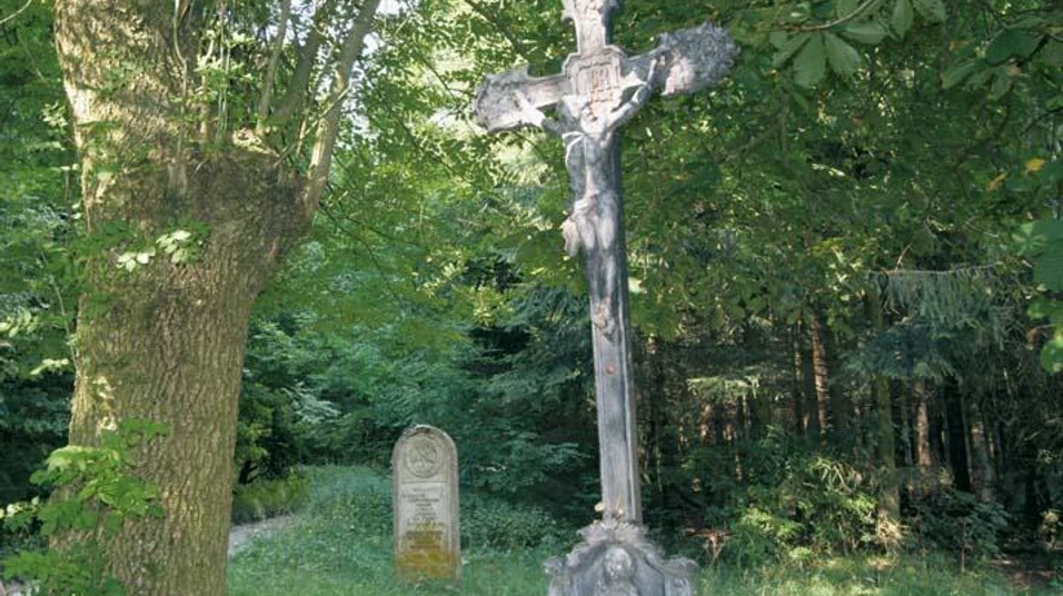 Kruzifix im Lippspringer Wald, umgeben von dichten Bäumen, mit einem Grabstein im Hintergrund.