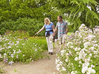 Skulpturenpark Lengerich - Jones Garten II Ein Paar spaziert auf einem Weg im Skulpturenpark, umgeben von blühenden Blumen und grünen Bäumen.