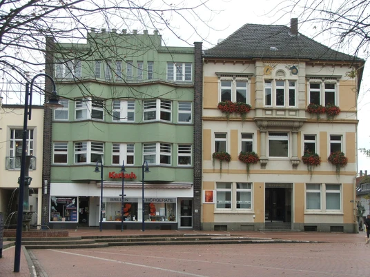 Zwei historische Gebäude nebeneinander, das linke in Pastellgrün, das rechte in Sandfarben gehalten.