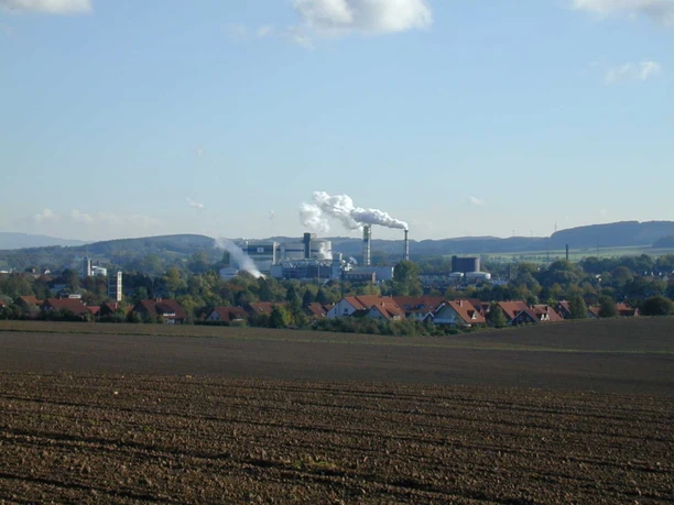 Zuckerfabrik Weitläufige Felder im Vordergrund, dahinter ein Industriegebäude mit rauchenden Schornsteinen.