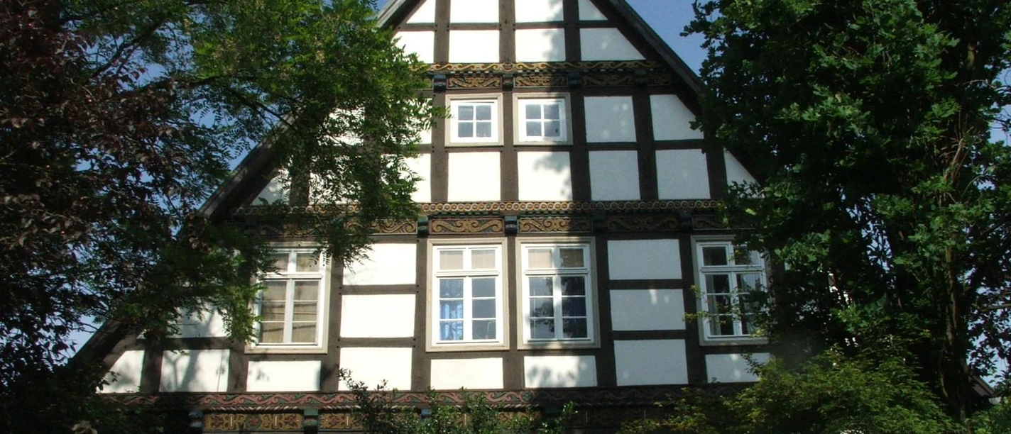 Fachwerkhaus Seppmann Fachwerkhaus Seppmann mit markantem Giebeldach, umgeben von üppig grünem Laubwerk bei Sonnenschein.