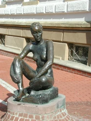Bronzestatue einer sitzenden Frau auf einem Sockel aus Ziegel in urbaner Umgebung.
