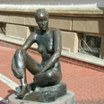 Bronzestatue einer sitzenden Frau auf einem Sockel aus Ziegel in urbaner Umgebung.