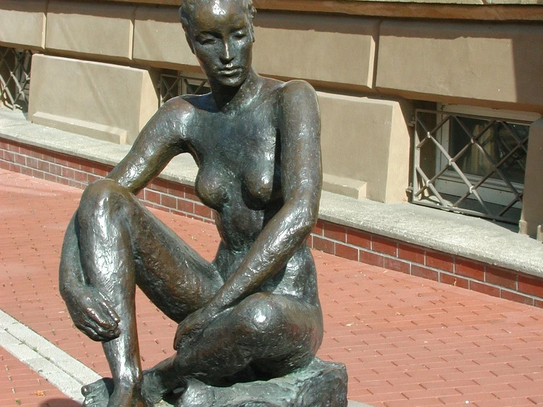 Große Sitzende Bronzestatue einer sitzenden Frau auf einem Sockel aus Ziegel in urbaner Umgebung.