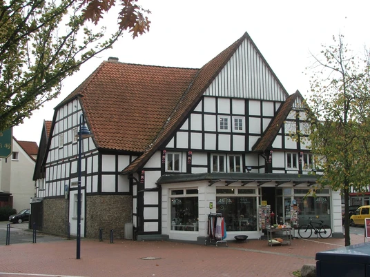 Fachwerkhaus mit rotem Ziegeldach und Geschäften im Erdgeschoss, umgeben von Laubbäumen.