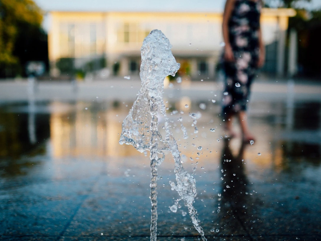Konzerthallenvorplatz Ein Springbrunnen spritzt Wasser in die Luft, im Hintergrund ist ein unscharfer Gebäudeeingang zu sehen.