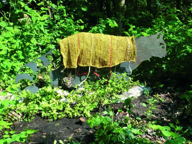 Des Katers neue Kleider Eisenkater-Skulptur im Grünen trägt gestrickten Umhang mit gelben und roten Streifen auf dem Rücken.