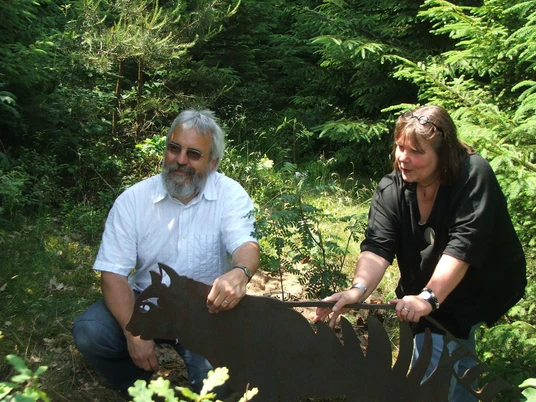 Stefanie und Wolfgang Pierel Stefanie und Wolfgang Pierel betrachten eine Metallskulptur eines Drachen im Wald. Sie wirken konzentriert.