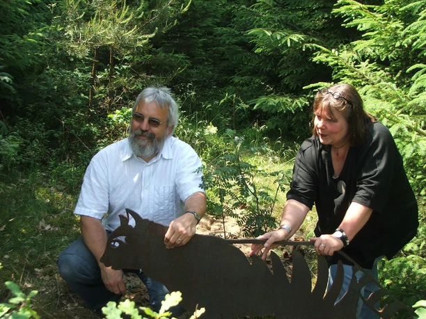 Stefanie und Wolfgang Pierel Stefanie und Wolfgang Pierel betrachten eine Metallskulptur eines Drachen im Wald. Sie wirken konzentriert.