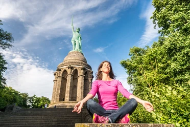Frau in rosa Pullover meditiert vor dem Hermannsdenkmal auf steinernen Stufen, Himmel blau.
