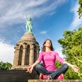 Frau in rosa Pullover meditiert vor dem Hermannsdenkmal auf steinernen Stufen, Himmel blau.