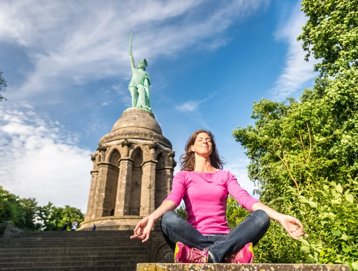 Frau in rosa Pullover meditiert vor dem Hermannsdenkmal auf steinernen Stufen, Himmel blau.