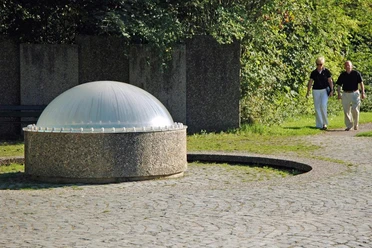 Gustav-Horstmann-Sprudel Ein Brunnen mit plastischer Kuppel auf einem gepflasterten Platz, umgeben von grüner Vegetation.