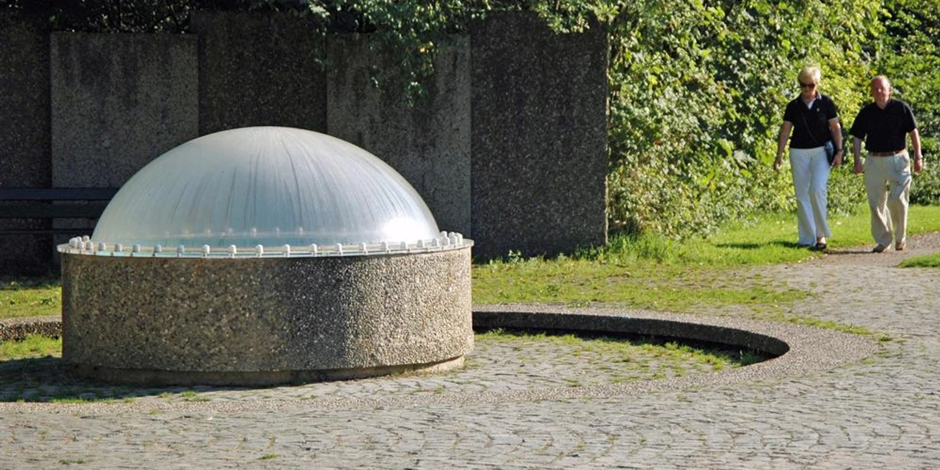 Gustav-Horstmann-Sprudel Ein Brunnen mit plastischer Kuppel auf einem gepflasterten Platz, umgeben von grüner Vegetation.