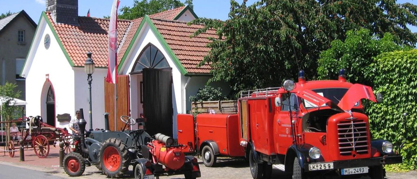 Historische Feuerwehrfahrzeuge und Traktoren parken vor einem kleinen Museum mit rotem Ziegeldach.
