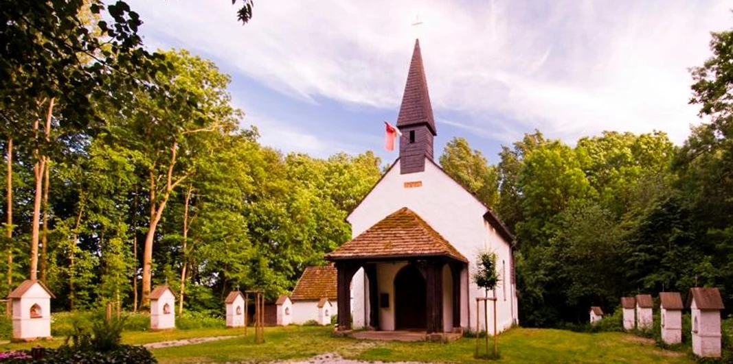 Eine kleine, weiße Kapelle mit dunklem Dach und Turm, umgeben von Bäumen und mehreren kleinen Stelen.