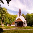 Eine kleine, weiße Kapelle mit dunklem Dach und Turm, umgeben von Bäumen und mehreren kleinen Stelen.