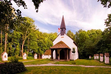 Klus Eddessen in Borgentreich-Borgholz Eine kleine, weiße Kapelle mit dunklem Dach und Turm, umgeben von Bäumen und mehreren kleinen Stelen.