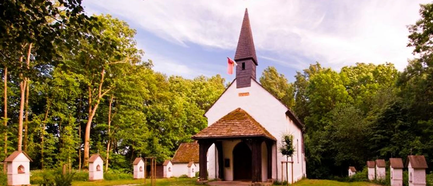 Klus Eddessen in Borgentreich-Borgholz Eine kleine, weiße Kapelle mit dunklem Dach und Turm, umgeben von Bäumen und mehreren kleinen Stelen.