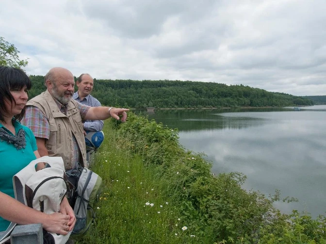 Drei Personen betrachten von einem bewachsenen Ufer aus einen ruhigen, von Wäldern umrahmten Stausee.