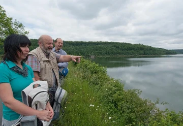 Aabach-Talsperre Drei Personen betrachten von einem bewachsenen Ufer aus einen ruhigen, von Wäldern umrahmten Stausee.