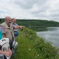 Aabach-Talsperre Drei Personen betrachten von einem bewachsenen Ufer aus einen ruhigen, von Wäldern umrahmten Stausee.