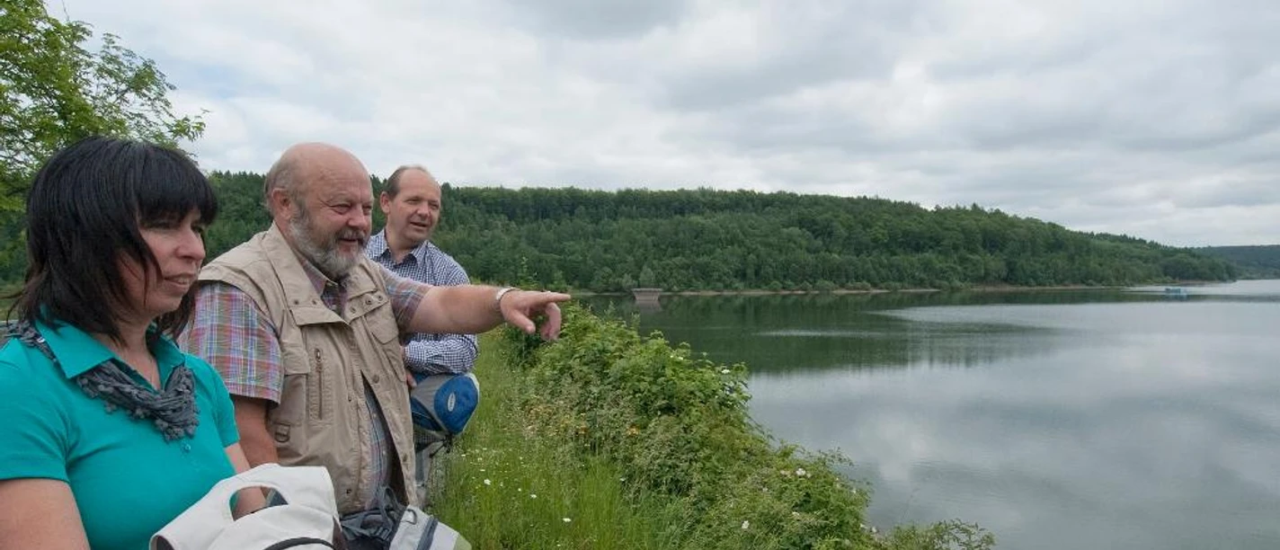 Aabach-Talsperre Drei Personen betrachten von einem bewachsenen Ufer aus einen ruhigen, von Wäldern umrahmten Stausee.