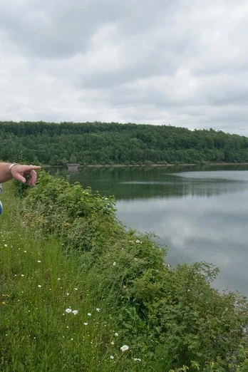 Aabach-Talsperre Drei Personen betrachten von einem bewachsenen Ufer aus einen ruhigen, von Wäldern umrahmten Stausee.