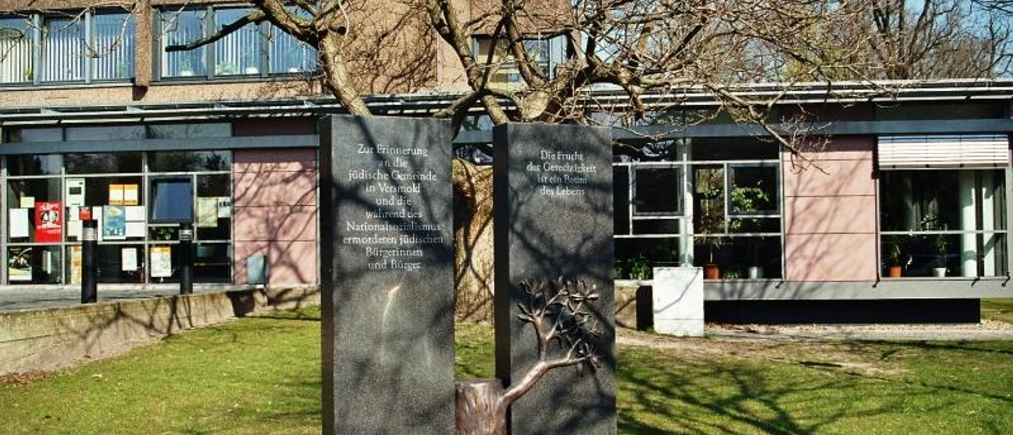 Erinnerungszeichen an die jüdische Gemeinde in Versmold Zwei Gedenktafeln neben einem Baum im Sonnenschein vor einem Gebäude, umrahmt von grüner Wiese.