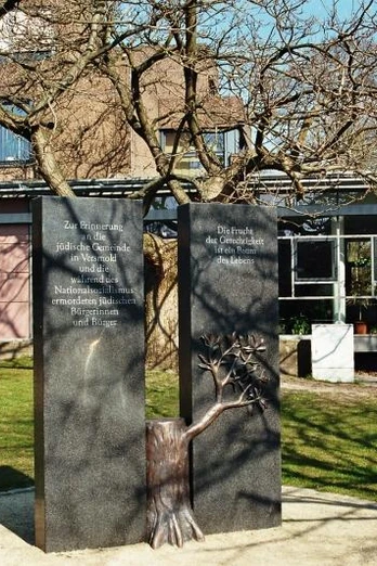Erinnerungszeichen an die jüdische Gemeinde in Versmold Zwei Gedenktafeln neben einem Baum im Sonnenschein vor einem Gebäude, umrahmt von grüner Wiese.