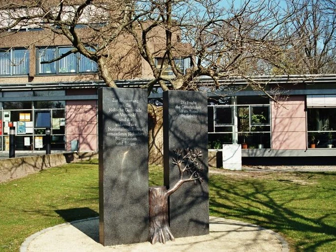 Erinnerungszeichen an die jüdische Gemeinde in Versmold Zwei Gedenktafeln neben einem Baum im Sonnenschein vor einem Gebäude, umrahmt von grüner Wiese.