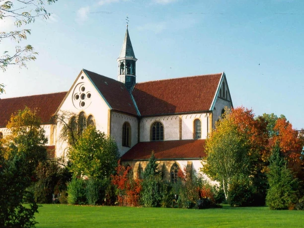 Ehemaliges Kloster Marienfeld: Abteikirche Kirche mit rotem Ziegeldach, umgeben von herbstlichen Bäumen und einem blauen Himmel im Hintergrund.