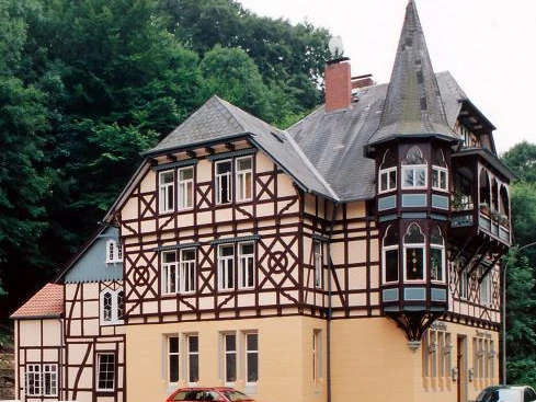 Historisches Fachwerkhaus mit drei Stockwerken und einem markanten spitzen Turm in bewaldeter Umgebung.