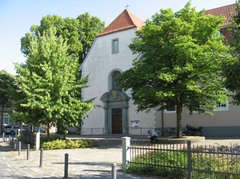 Eine kleine Kirche mit rotem Ziegeldach, flankiert von grünen Bäumen, und einem ruhigen Vorplatz.
