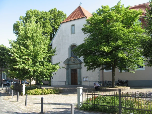 Klosterkirche Eine kleine Kirche mit rotem Ziegeldach, flankiert von grünen Bäumen, und einem ruhigen Vorplatz.