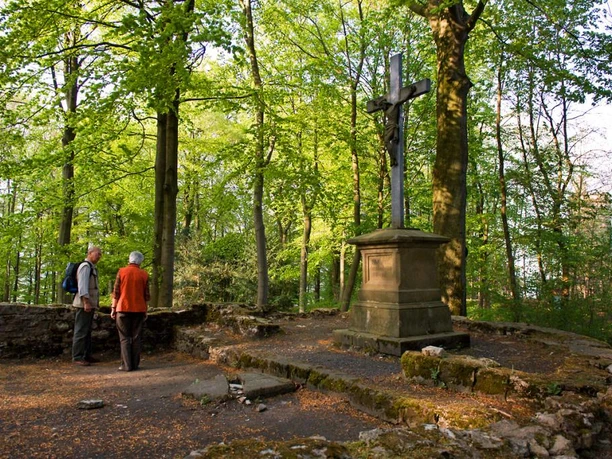 Kreuz an der Iburg Ein hölzernes Kreuz auf einem steinernen Podest im lichten Wald, umgeben von zwei Menschen.