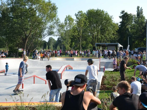 Menschen unterschiedlicher Altersgruppen genießen einen sonnigen Tag im Skatepark mit Grünflächen und Bäumen.