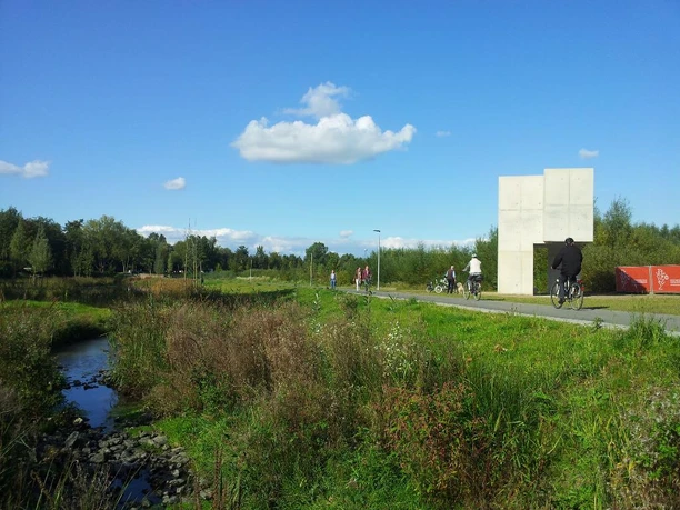 "Pavillon" by Christian Odzuck Radfahrer und Spaziergänger auf einem sonnigen Weg in einem grünen, naturnahen Park mit modernem Bauwerk.