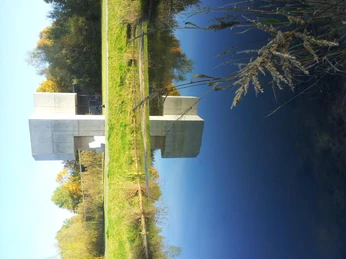 "Pavillon" Modernes, graues Gebäude mit rechteckigen Elementen, steht an einem ruhigen, reflektierenden Gewässer.